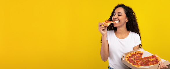 A woman with curly hair takes a bite of pizza while holding a pizza box. She smiles happily,...