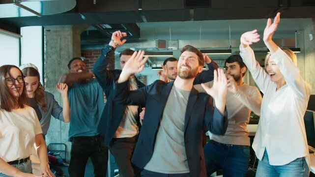 Group of coworkers celebrating success. Big team dancing with raised hands and laughing together. People enjoying shared achievement in energetic moment. In bright modern office location.