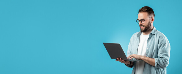 A young man stands against a bright blue backdrop, focused on his laptop. He wears glasses and a casual outfit, appearing engaged and cheerful while typing.
