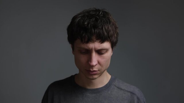 Anxious man looking into camera then lowering gaze. Portrait of stress, worry and uncertainty on neutral background.