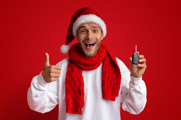 A joyful man wearing a Santa hat and a red scarf smiles widely while giving a thumbs up. He holds a car key and stands against a bright red background, capturing the festive spirit of Christmas.