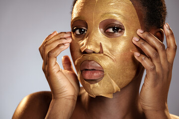 Woman applying a gold facial mask, showcasing skincare routine and self-care on a neutral background