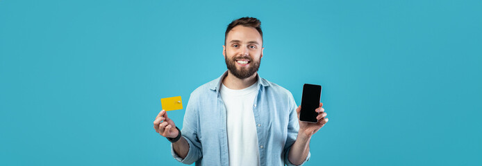A man with a beard smiles while standing against a bright blue background, holding a smartphone in one hand and a credit card in the other. He appears enthusiastic about digital payments.