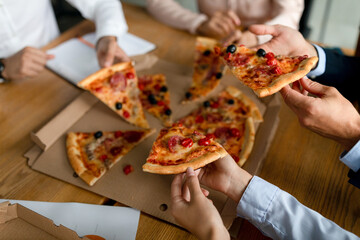 Colleagues gather around a box of pizza during their lunch break in the office. They happily take slices, enjoying a tasty meal together while sitting at their desks.