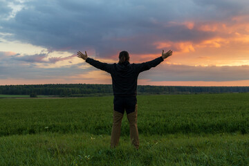 A man stands in a field with his arms outstretched, looking up at the sky