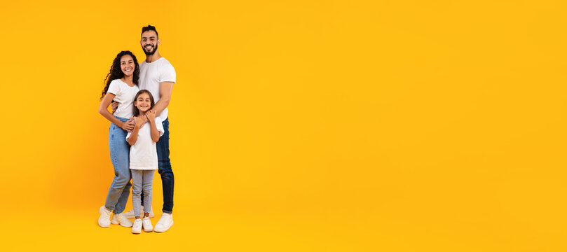 A cheerful family stands close together against a vibrant yellow backdrop. The parents are smiling, embracing their daughter who stands in front. They all wear casual white tops and jeans.