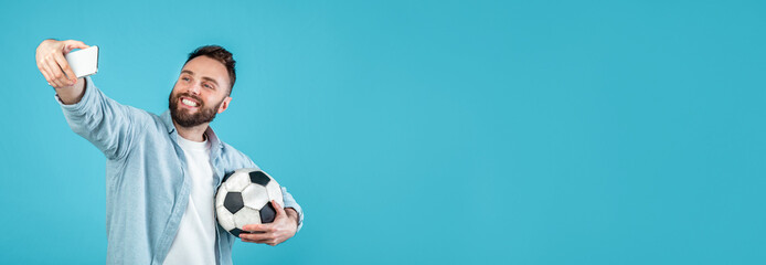 A young man is smiling as he takes a selfie with a soccer ball under his arm. The bright blue...