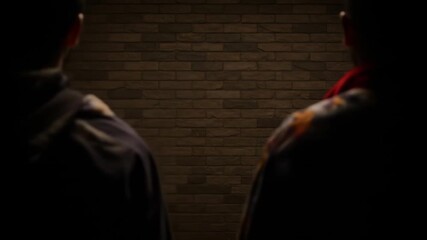 Two figures stand silhouetted against a dark, rough brick wall, creating a mysterious and possibly suspenseful atmosphere