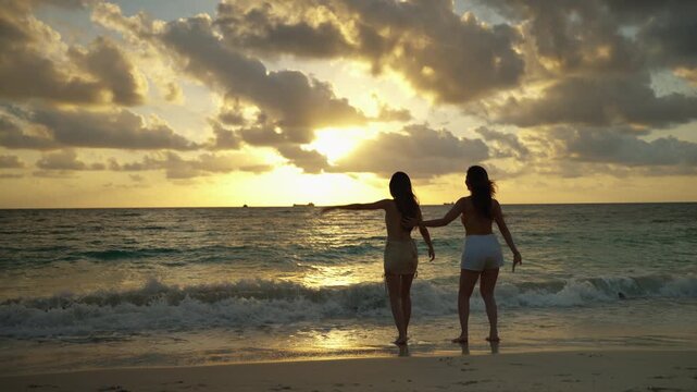 Two women stand together at the shoreline during a dramatic sunset, enjoying the warm golden light and gentle waves. Their playful, relaxed pose reflects freedom, friendship, and appreciation of natur