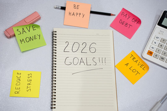 Notebook with 2026 goals written on it. Several sticky notes surround it, listing goals like save money and pay off debt. New year resolutions for 2026 year.