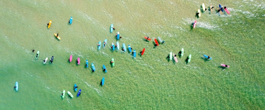 Aerial view of surfers dotting the turquoise waters of the beach, their colorful boards a vibrant contrast against the sandy shore, Newquay, England, United Kingdom.