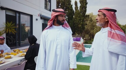 Two arab men, senior and younger, in traditional thobes and keffiyehs discuss property options by a modern house with pool, elder pointing and guiding son on investment decisions