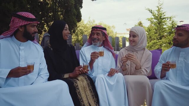 Five joyful middle eastern muslim family members and friends, dressed in traditional clothing, sitting together enjoying tea and a snack outdoors in a garden, celebrating a cultural gathering
