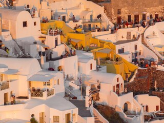 Aerial view of white buildings cascade down the hillside under a warm glow, contrasting with the deep shadows and vibrant hues of Oia, Thira, Greece.