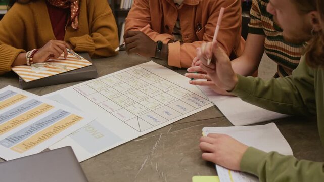 High angle shot of male teacher explaining English grammar rule to diverse group of adult students pointing at printout with irregular verbs while sitting around library table