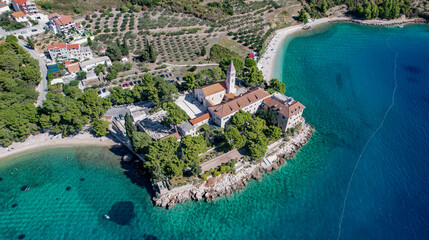 Aerial view of a historic monastery on a rocky peninsula surrounded by the turquoise Adriatic Sea, framed by lush green trees and distant buildings, Bol, Split-Dalmatia County, Croatia.