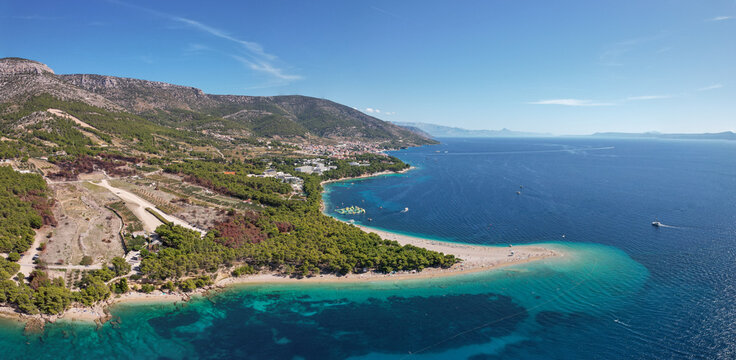 Aerial view of Zlatni Rat's iconic golden sand beach meeting the turquoise Adriatic, backed by lush green hills and a clear blue sky, Bol, Split-Dalmatia County, Croatia.