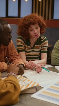 Vertical shot of young female teacher pointing at printout table of English irregular verbs explaining grammar rule to diverse group of students gathered in library