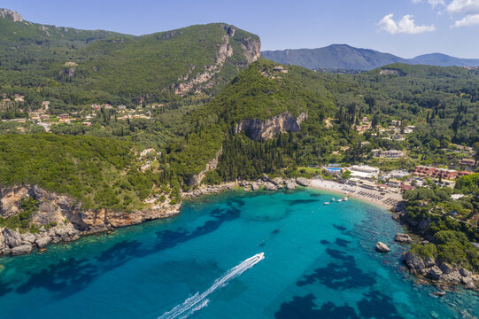 Aerial view of a boat cutting through the turquoise waters near a beach framed by rugged cliffs and verdant hillsides, Corfu, Corfu, Greece.