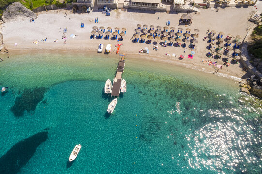 Aerial view of boats bobbing near a wooden pier extending onto the beach, framed by neat rows of umbrellas against the turquoise waters, Corfu, Corfu, Greece.