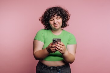A person with curly hair in a green shirt using a smartphone against a pink background.