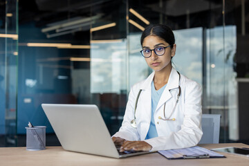 Portrait of a serious young Indian female doctor in a white coat sitting at an office desk in a hospital, working on a laptop and looking at the camera