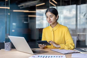 Serious young Indian businesswoman sitting in the office at a table with documents and a laptop and using a calculator