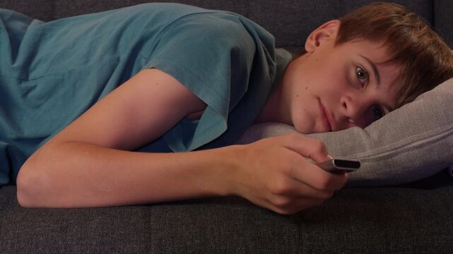 Teenager boy reclining on couch with remote control in hand, flipping through channels and appearing disinterested before eventually dozing off from exhaustion in evening at residence
