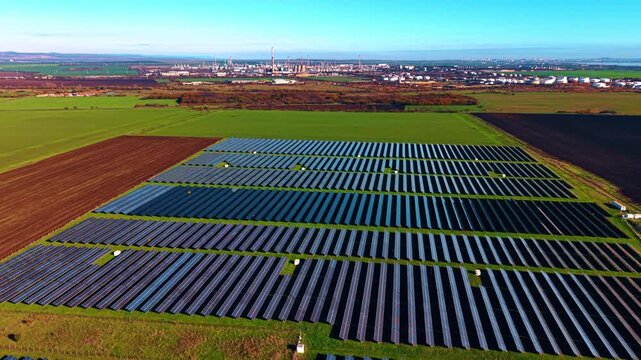 Countryside with large fields of photovoltaic cells used for production renewable energy. Green power concept. Oil refinery at backdrop.