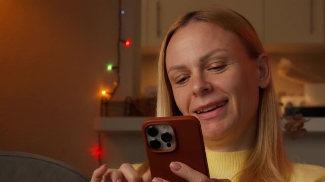 Happy woman reacting with joy upon receiving exciting news on phone, expression shifting from smile to tears of happiness as she sits at home with colorful string lights in background