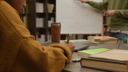 Close up on hand of multiethnic young woman writing notes in notebook during group Spanish class in library