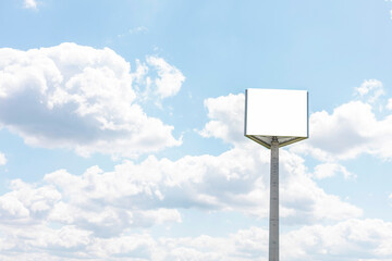 The signboard is empty and white, rising on a pole in an open space. The sky is filled with clouds,...