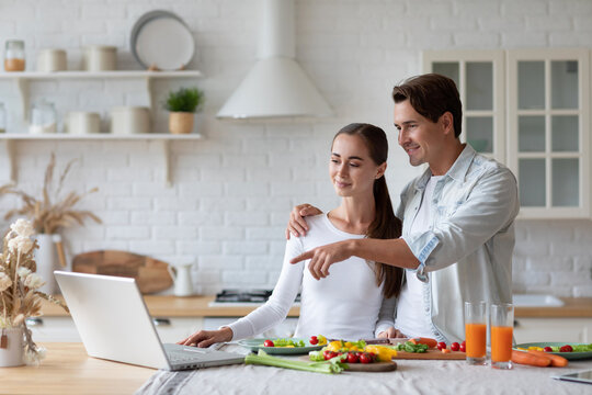Woman in a white long-sleeve shirt pointing at a laptop, standing in a kitchen with a man in a light blue shirt behind her.