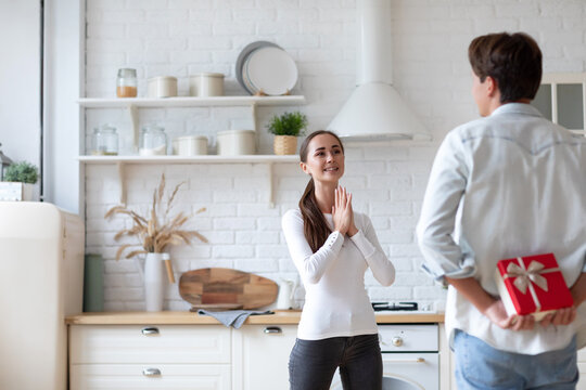 Woman in a white long-sleeve shirt clapping her hands, standing in a kitchen with a man in a light blue shirt behind her. The man is holding a red gift box with a white ribbon.