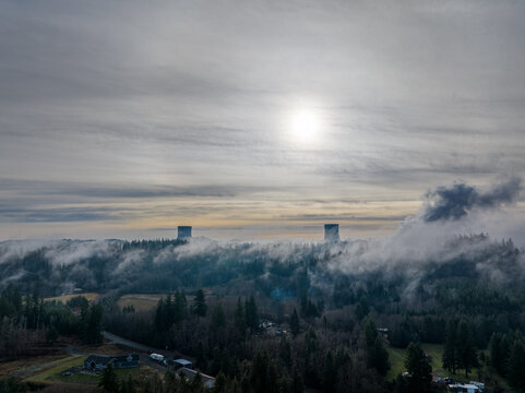 Aerial view of twin cooling towers emerge through the dense fog, piercing the skyline above verdant forest, Elma, Washington, United States.