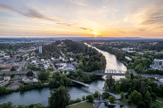 Aerial view of the Willamette River winds through the city, reflecting the warm hues of the setting sun, with Skinner Butte Park rising majestically in the distance, Eugene, Oregon, United States.