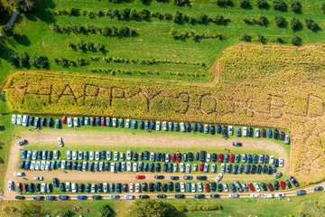 Aerial view of a vast corn maze with 'HAPPY 30TH BDAY' carved into its golden expanse, juxtaposed against rows of parked cars under the clear sky, Peekskill, New York, United States.