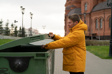 A man opens a green dumpster while standing in a city park on a cloudy day