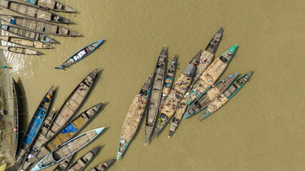 Aerial view of wooden boats clustered on the tranquil river, carrying goods under the bright sunlight, creating a vibrant scene, Kazipur, Rajshahi Division, Bangladesh.