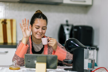 Young woman sits at home, talking on a video call on her tablet while sipping her morning coffee.