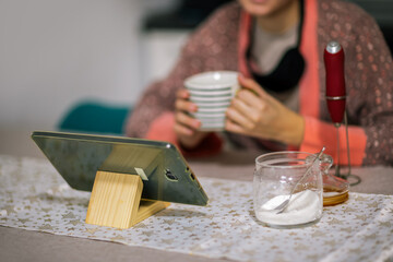 Young woman sits at home, looking at her tablet while sipping her morning coffee.
