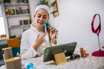 Young woman sits at home, filming a beauty vlog on her tablet and giving skincare and makeup advice.