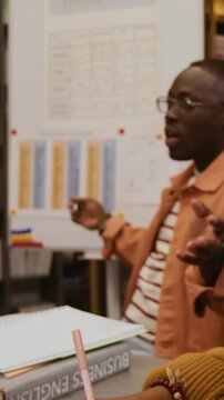 Vertical shot of African American male teacher conducting business English class explaining grammar rules on whiteboard while female student writing notes in notebook studying in library