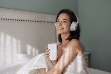 Young woman with headphones enjoying a warm beverage while sitting in bed, surrounded by soft bedding and a calming atmosphere, embracing a cozy morning routine