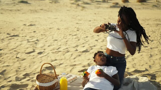 African american mother with braids capturing special moments of son on sunny beach using vintage film camera during family picnic, creating precious summer memories