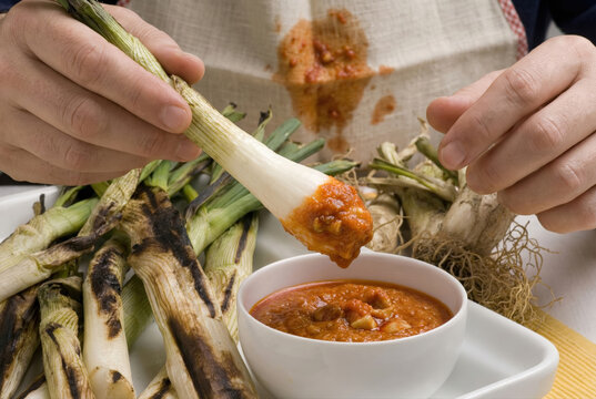 Primer plano de cal&ccedil;ot asado tradicional siendo sumergido en salsa romesco durante una cal&ccedil;otada, con babero manchado, manos sosteniendo el cal&ccedil;ot y un fondo de cal&ccedil;ots asados listos para comer.
