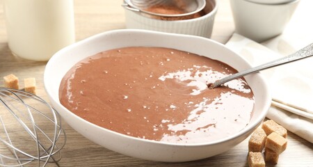 Liquid chocolate dough in bowl, milk, cocoa powder, brown sugar and whisk on wooden table, closeup