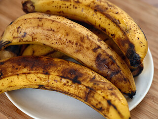 A bunch of overripe bananas with brown spots resting on a white plate on a wooden table. Close up.
