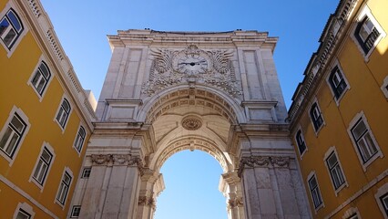 Elaborate stone archway under blue sky, flanked by yellow buildings, centered