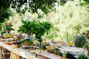 Wide Angle Shot of a Long Rustic Wooden Outdoor Buffet Table Laden with Diverse Gourmet Dishes, Salads, Breads, and Fresh Herbs in a Lush Garden Setting